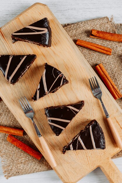 Rebanadas de pastel de chocolate en una tabla de madera y saco con canela y tenedores vista de ángulo alto sobre un fondo blanco.