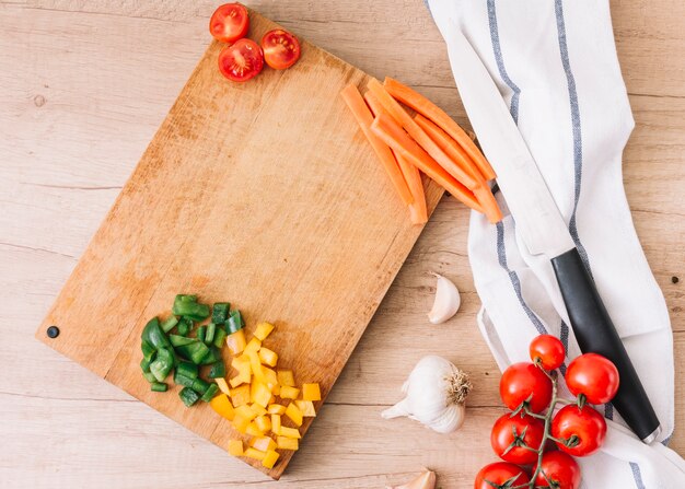 Rebanadas de pimientos; Zanahoria; tomates a la mitad en la tabla de cortar con bulbo de ajo; Cuchillo y servilleta sobre el escritorio de madera.
