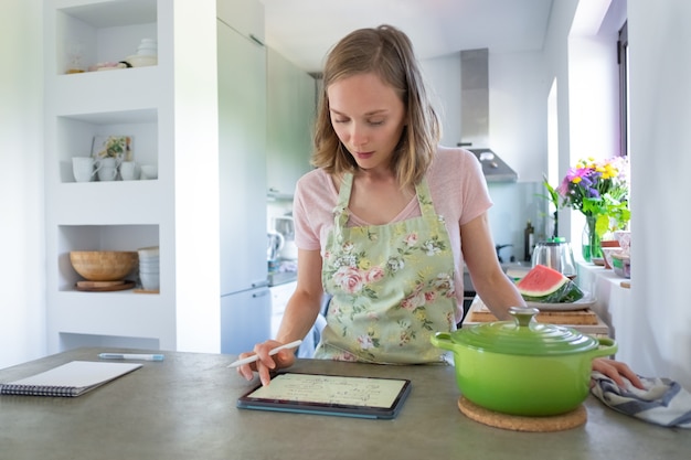 Receta de consultoría joven enfocada mientras cocina en su cocina, usando tableta cerca de una cacerola grande en el mostrador. Vista frontal. Cocinar en casa e internet concepto