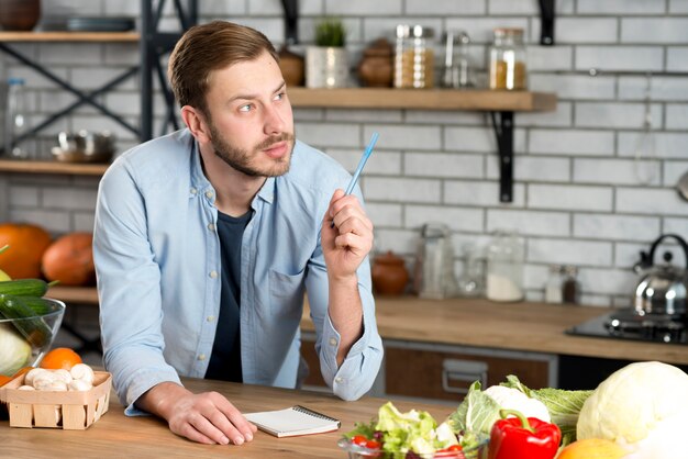 Receta pensativa joven de la escritura del hombre en cocina con la pluma y el diario