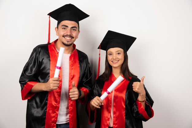 Recién graduados con diploma haciendo pulgares hacia arriba en blanco.