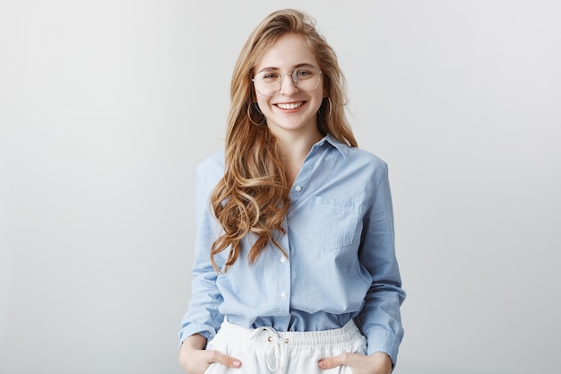 Los recién llegados conocen a sus compañeros de equipo. Agradable empresaria guapa en gafas transparentes con camisa de cuello azul, sonriendo ampliamente, asegurando la buena calidad del producto sobre la pared gris