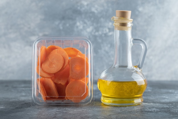 Recipiente de plástico lleno con rodajas de zanahoria y botella de aceite sobre fondo gris.