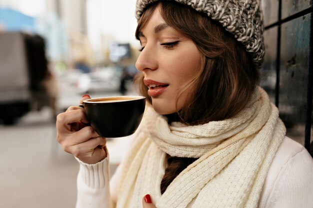 Recorta el retrato exterior de una linda chica encantadora con gorro de punto y bufanda bebiendo café y disfrutando de una pausa para el café