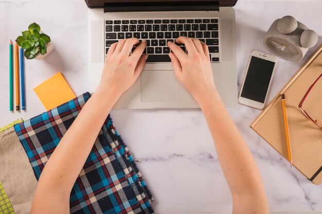 Recortar las manos escribiendo en la computadora portátil cerca de muestras de tela