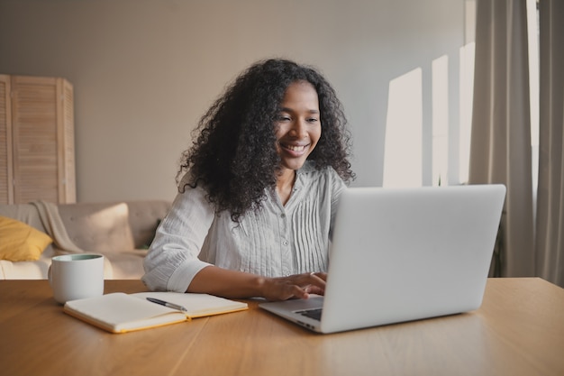 Redactor alegre joven afroamericana sentado frente a la computadora portátil abierta con taza y cuaderno en el escritorio, sintiéndose inspirado, trabajando en un nuevo artículo de motivación. Personas, ocupación y creatividad