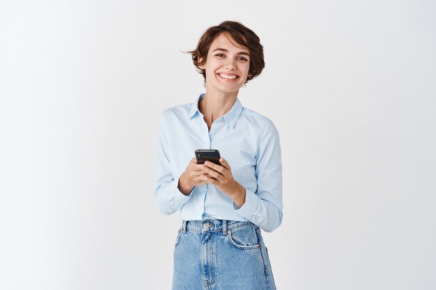 Redes. Sonriente mujer profesional sosteniendo smartphone de pie en la pared blanca