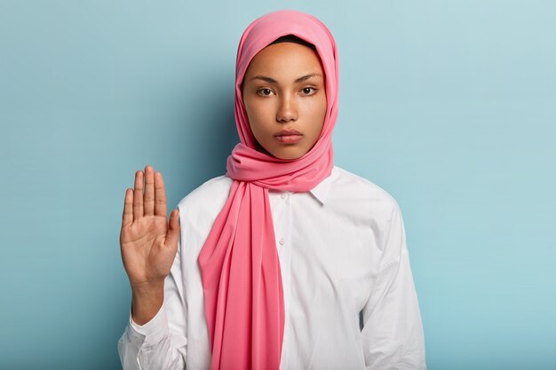 Reduzca la velocidad y pare. Grave mujer árabe de piel oscura muestra un gesto de rechazo, levanta la palma hacia, expresa su rechazo, usa un sombrero rosa y una camisa blanca, aislada sobre una pared azul.