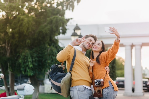 Refinada mujer de cabello oscuro haciendo selfie con hombre guapo en camisa naranja con arquitectura blanca sobre fondo