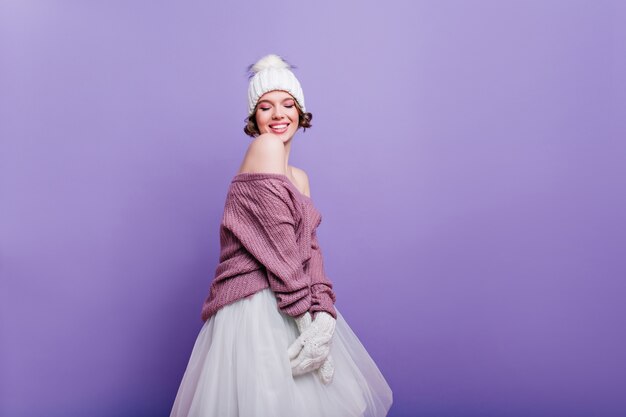 Refinada mujer europea con sombrero blanco y falda posando con expresión facial inspirada. chica de pelo corto de moda en suéter aislado en la pared púrpura.