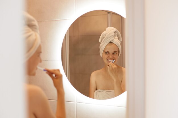 Reflejo en el espejo de una atractiva joven adulta con una toalla blanca en el pelo posando en el baño y cepillándose los dientes, procedimientos de higiene después de tomar una ducha.