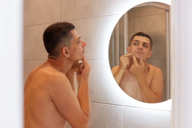 Reflexión en el espejo guapo hombre de pelo oscuro de pie con la parte superior del cuerpo desnudo y mirando su rostro, encuentra granos, problemas de piel, procedimientos de higiene matutina.