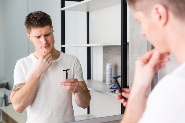 Reflexión del hombre joven serio con la mano en su barbilla que mira la maquinilla de afeitar en espejo