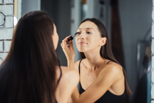 Reflexión de la joven y bella mujer aplicando su maquillaje, mirando en un espejo