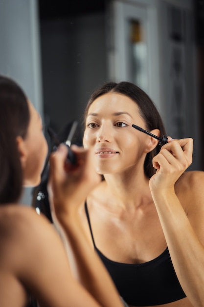 Reflexión de la joven y bella mujer aplicando su maquillaje, mirándose en un espejo