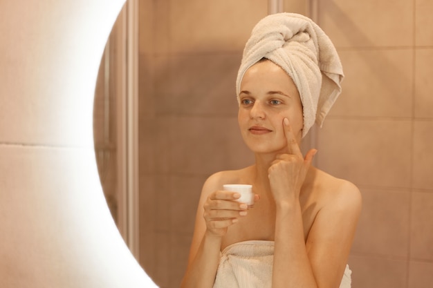 Reflexión de una mujer adulta joven en el espejo aplicando crema cosmética en la cara, poniéndose argent nutritivo en su piel facial en el baño, posando con una toalla en la cabeza.