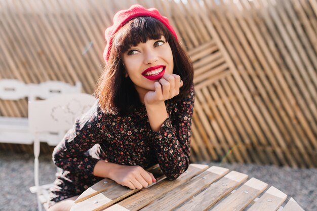 Reflexiva chica francesa elegante espera el café de la mañana, sentada en la mesa de madera en un café de la calle. Retrato de mujer joven soñadora con peinado corto con boina roja de moda y vestido vintage