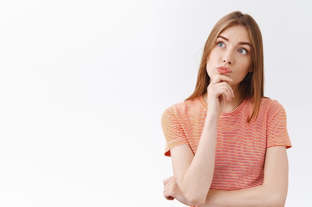 Reflexiva e intrigada joven rubia con camiseta a rayas, tocar la barbilla y hacer pucheros con curiosidad, mirar la esquina superior izquierda intrigada e insegura, tomar una decisión, pensar en una decisión, fondo blanco