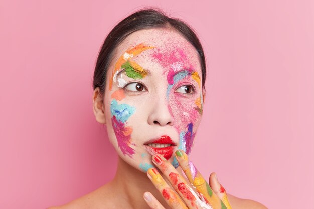 Reflexiva hermosa mujer asiática manchada con pinturas de acuarelas brillantes trabaja como artista se encuentra sin camisa aislado sobre fondo rosa