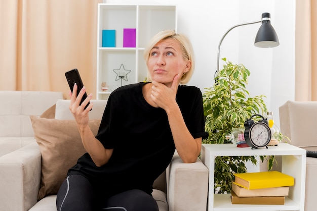 Reflexiva hermosa mujer rusa rubia sentada en un sillón poniendo la mano en la barbilla y sosteniendo el teléfono mirando hacia arriba dentro de la sala de estar