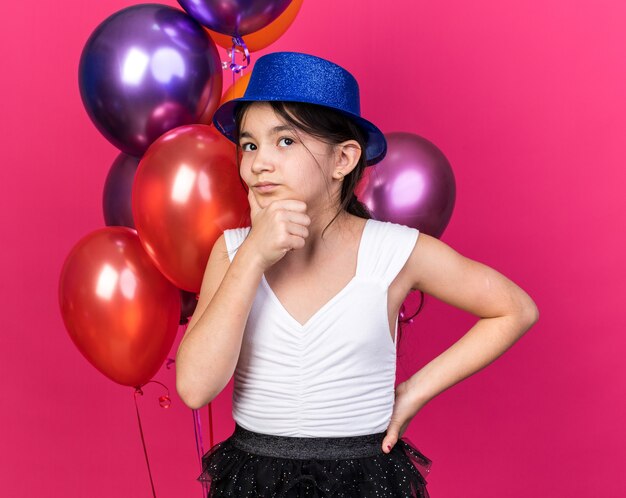 Reflexiva joven caucásica con gorro de fiesta azul sosteniendo la barbilla de pie delante de globos de helio mirando hacia arriba aislado en la pared rosa con espacio de copia