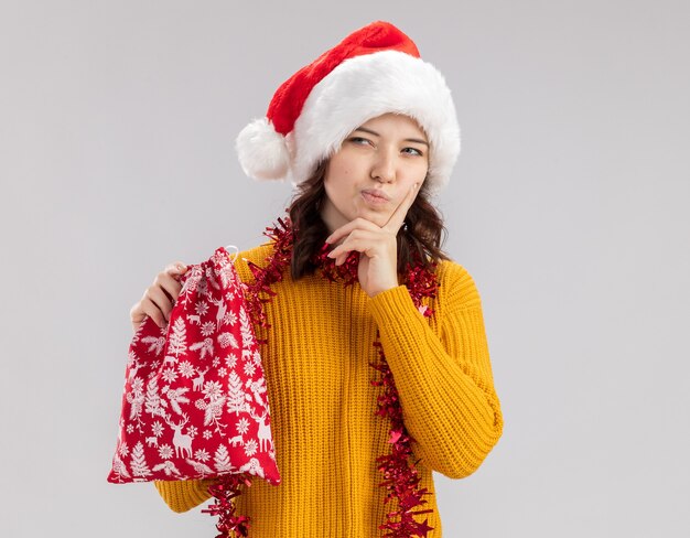 Reflexiva joven eslava con gorro de Papá Noel y con guirnalda alrededor del cuello pone el dedo en la barbilla y sostiene la bolsa de regalo de Navidad mirando al lado aislado en la pared blanca con espacio de copia