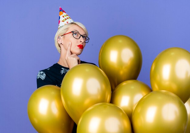 Reflexiva joven rubia fiestera con gafas y gorra de cumpleaños de pie detrás de globos tocando la cara y mirando hacia arriba aislado sobre fondo púrpura