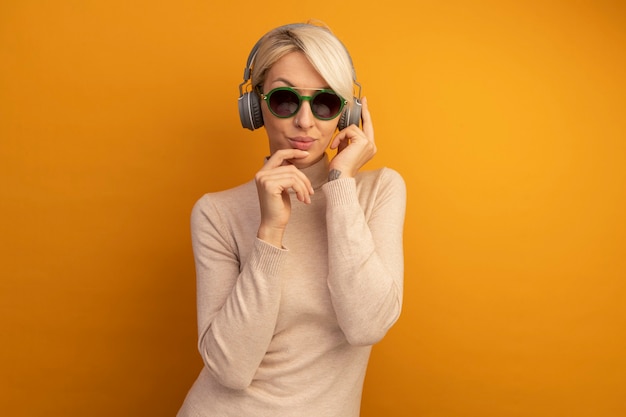 Reflexiva joven rubia con gafas de sol y auriculares agarrando auriculares tocando la barbilla