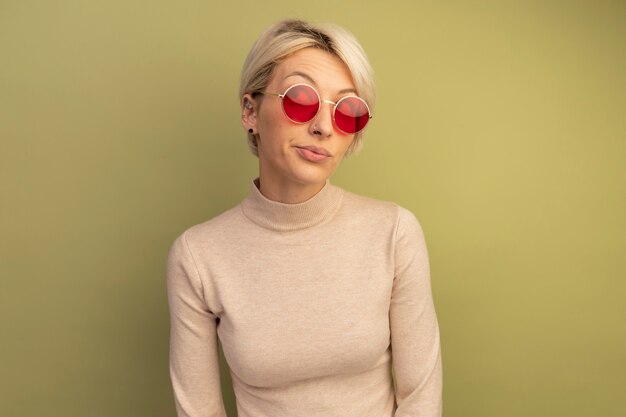 Reflexiva joven rubia con gafas de sol mirando al lado aislado en la pared verde oliva con espacio de copia