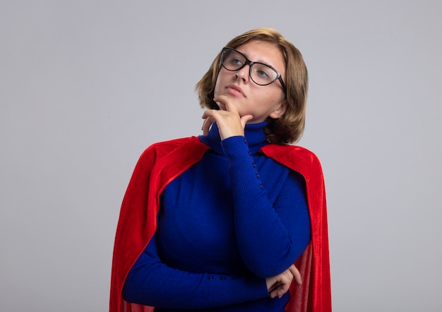 Reflexiva joven superhéroe rubia en capa roja con gafas tocando la barbilla mirando al lado aislado sobre fondo blanco.
