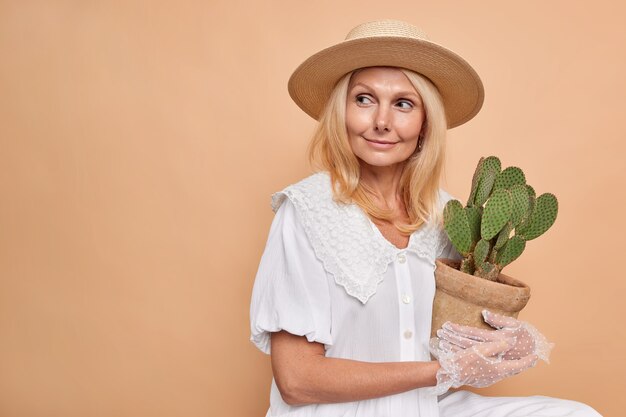 Reflexiva mujer aristocrática de mediana edad soñadora mira hacia otro lado comprado cactus en macetas para el jardín de su casa le gustan las plantas de interior usa un vestido fedora blanco de moda y guantes de encaje aislados sobre una pared beige