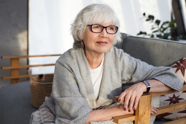 Reflexiva mujer madura de pelo gris de mediana edad con anteojos, bufanda ancha y reloj de pulsera pasar tiempo libre en casa, sentado en un cómodo sofá en la sala de estar, con mirada pensativa