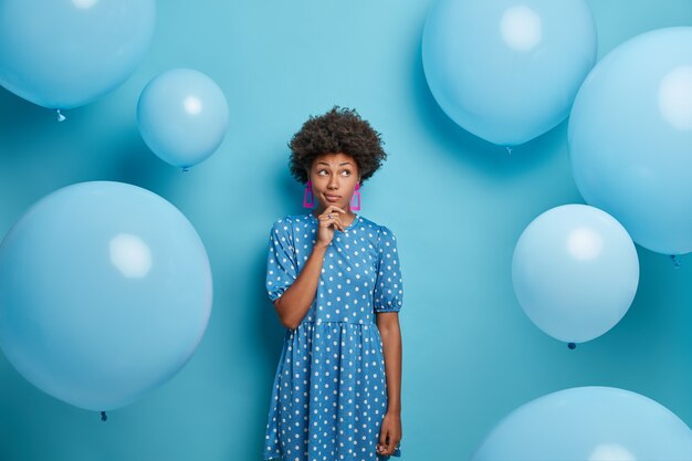 Reflexiva mujer de piel oscura usa un vestido azul de lunares, está en una fiesta de globos, vestida con un atuendo de moda, tiene una expresión pensativa, va a celebrar el cumpleaños. Tiempo libre, concepto de fiesta.