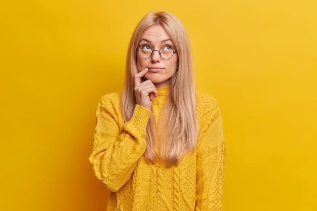 Reflexiva mujer rubia concentrada por encima de estar sumido en pensamientos lleva gafas redondas suéter casual