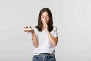 Foto gratuita reflexiva niña bonita sonriente reflexionando mientras sostiene el pedazo de pastel, blanco.