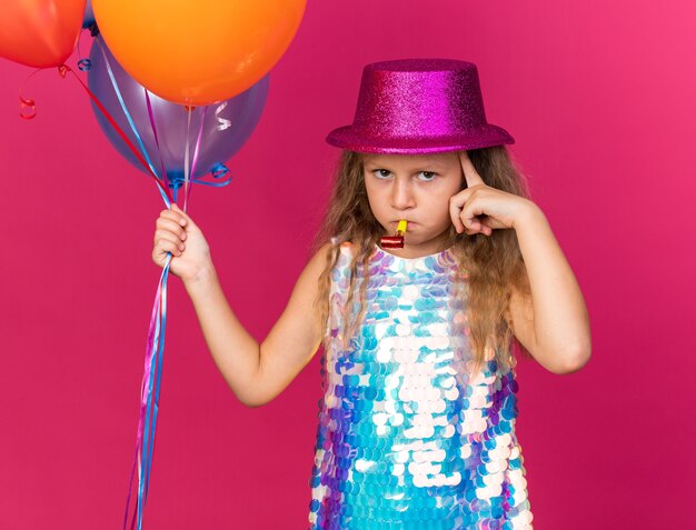 Reflexiva niña rubia con gorro de fiesta púrpura sosteniendo globos de helio y soplando silbato de fiesta aislado en la pared rosa con espacio de copia