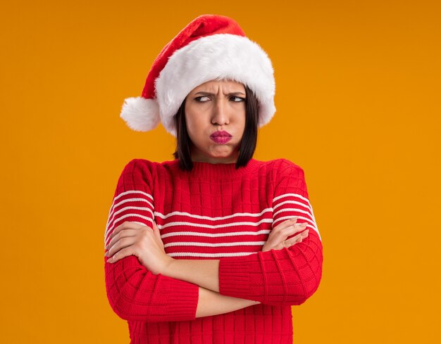 Refunfuñar joven vistiendo gorro de Papá Noel de pie con postura cerrada mirando al lado hinchando las mejillas aislado en la pared naranja con espacio de copia