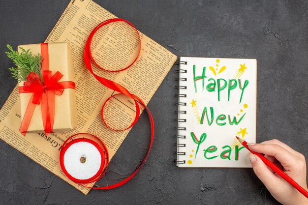 Regalo de Navidad de vista superior en cinta de abeto de rama de papel marrón en periódico feliz año nuevo escrito en lápiz de Bloc de notas en mano femenina sobre superficie oscura