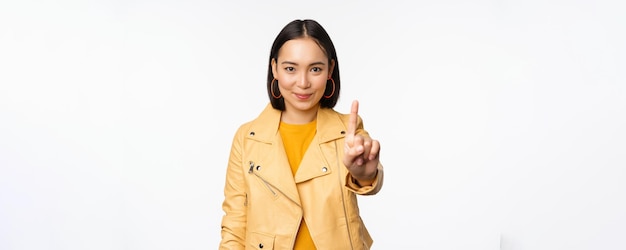 Regla número uno Mujer asiática sonriente segura mostrando el dedo índice detener prohibir el gesto desaprobar algo de pie sobre fondo blanco