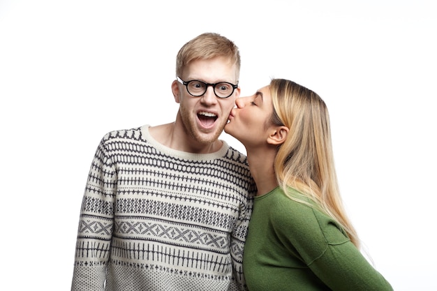 Regocijado emocional joven nerd masculino barbudo con anteojos exclamando con entusiasmo, sorprendido mientras hermosa mujer lo besa en la mejilla