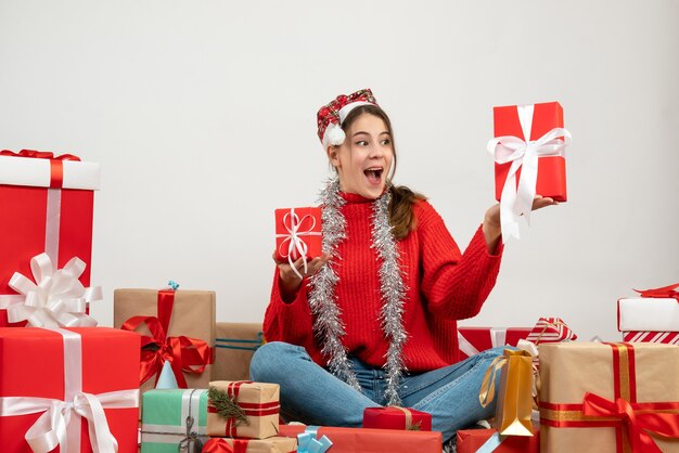 Regocijado niña con gorro de Papá Noel sosteniendo regalos sentados alrededor de regalos en blanco