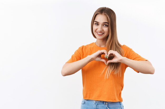 Relación, emociones y concepto de juventud. Retrato de chica rubia alegre y atractiva en camiseta naranja, muestra el signo del corazón sobre el pecho para expresar amor, cuidado y simpatía, apasionado por algo
