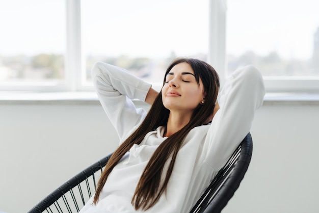 Foto gratuita relajación total mujer joven atractiva cogida de la mano detrás de la cabeza y sonriendo mientras está sentada en la silla en casa