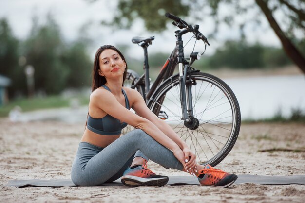 Relajado y satisfecho. Ciclista femenina con buena forma corporal sentada cerca de su bicicleta en la playa durante el día