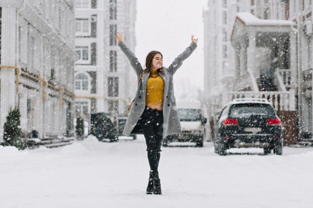 Relajante mujer caucásica posando con las manos en alto bajo la nieve en la calle urbana. Foto de cuerpo entero al aire libre de una mujer bonita en suéter amarillo y abrigo gris disfrutando el fin de semana en la ciudad de invierno.