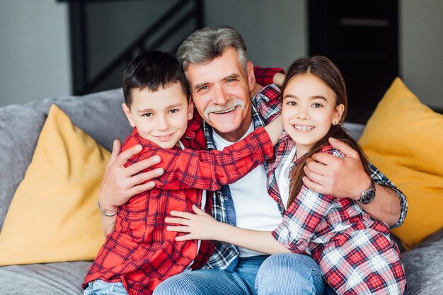 Relajarse. Abuelo alegre feliz sonriendo y sentado en el sofá con sus nietos y abrazando a sus.