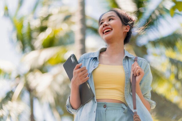 Relajarse casual despreocupado sonriente asiático femenino nómada digital independiente programador portátil de mano caminando en la playa y cocotero azul cielo nuevo estilo de vida milenario trabajando en cualquier lugar con felicidad