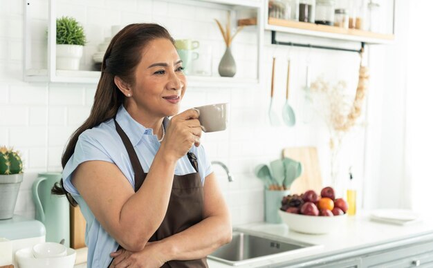 Relájese anciana asiática en delantal sosteniendo una taza de café en la cocina