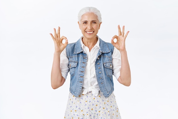 Relájese, una anciana feliz sin molestias con un corte de pelo gris, use un vestido de chaleco de mezclilla que muestre todo bien, ok, gesto sonriente, tenga las cosas bajo control, asegure todo el fondo blanco fino