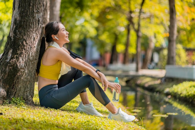 Relájese el tiempo de descanso de la deportista asiática casual después de terminar la mañana, hacer ejercicio, un estilo de vida saludable, la mujer asiática, relajarse, refrescarse después de correr, entrenar bajo el árbol cerca del agua en el parque